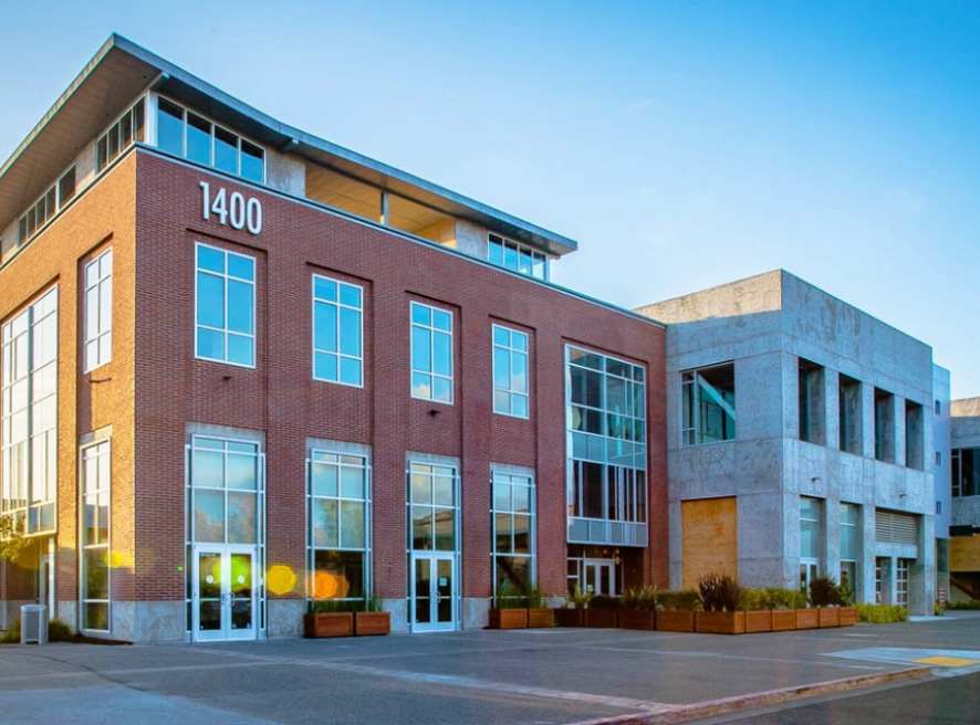 A modern brick and concrete building with large windows displaying the number 1400 at the top corner. The entrance is flanked by planters. The sky is clear and blue.