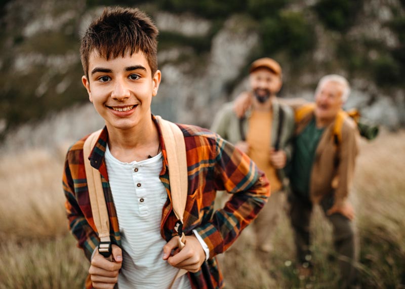 A young person in a plaid shirt and backpack stands in the foreground, smiling. Two adults are in the background, out of focus, in a grassy, hilly area.