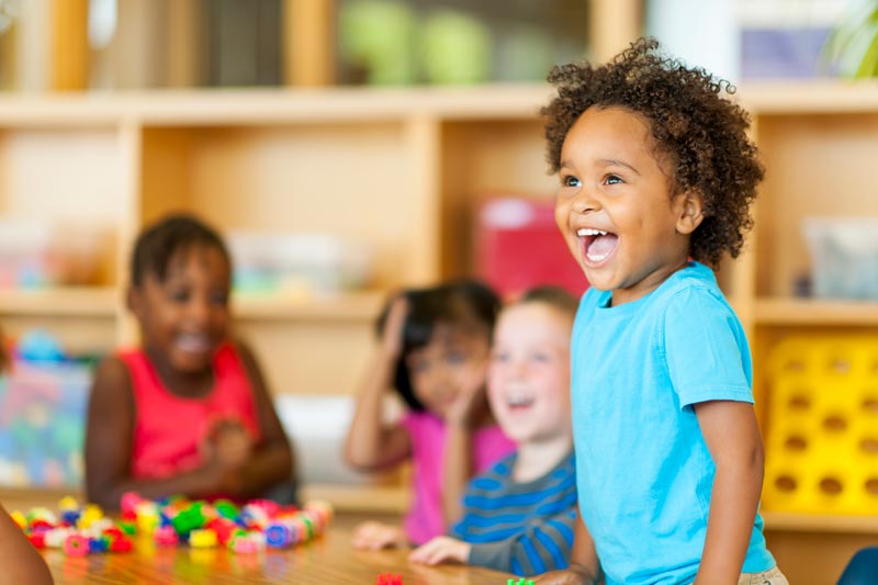 Children are smiling and playing with colorful building toys at a table in a classroom.