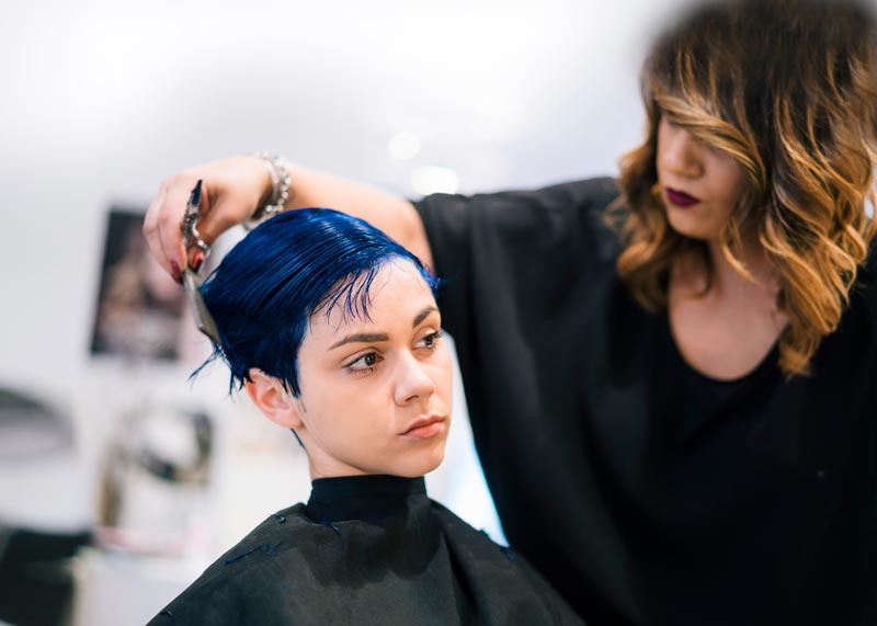 A stylist cuts a person's short, blue hair in a salon.