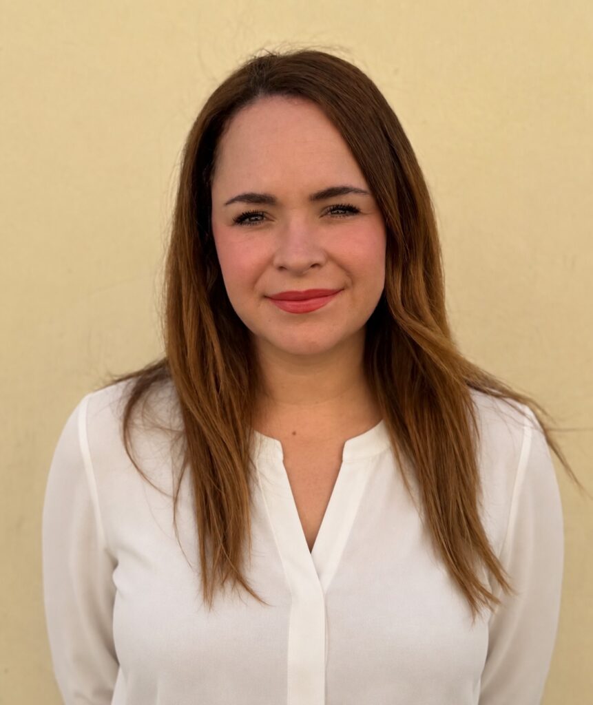 Woman with long brown hair wearing a white blouse stands in front of a plain light yellow wall, looking at the camera with a neutral expression.