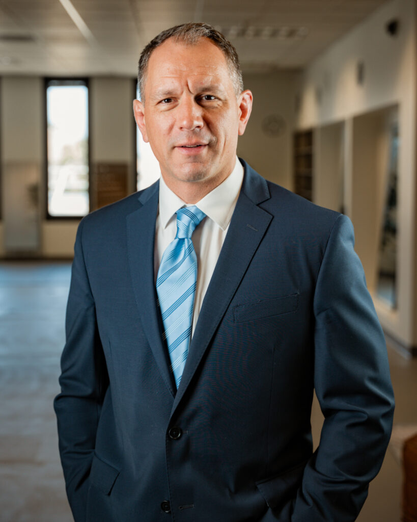 A man in a blue suit and striped tie stands indoors, looking at the camera with a neutral expression. The background shows a hallway with windows and neutral decor.