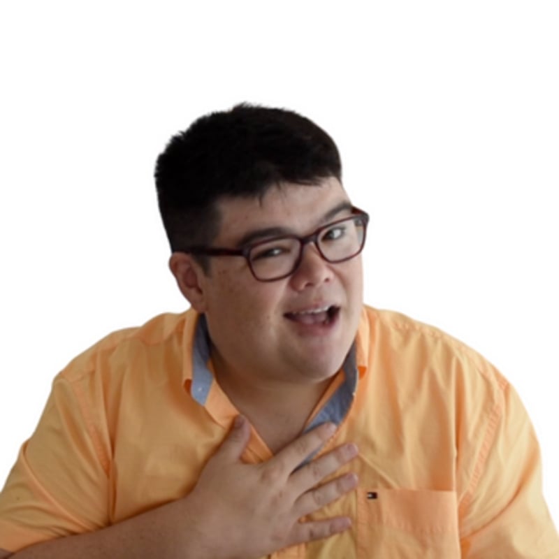A person wearing glasses and a light orange shirt smiles and gestures with one hand on their chest against a white background.