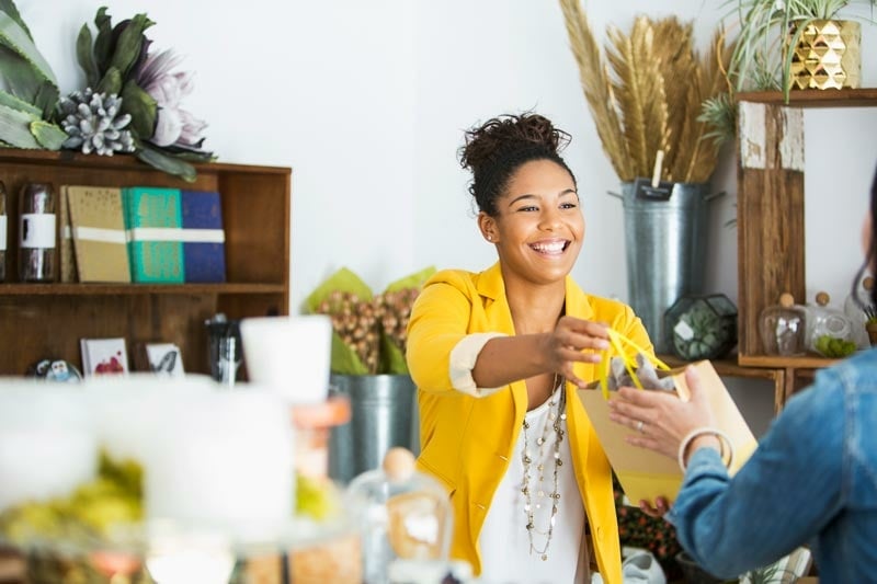 A smiling shop worker in a yellow blazer hands a paper shopping bag to a customer across a counter in a store decorated with plants and shelves.