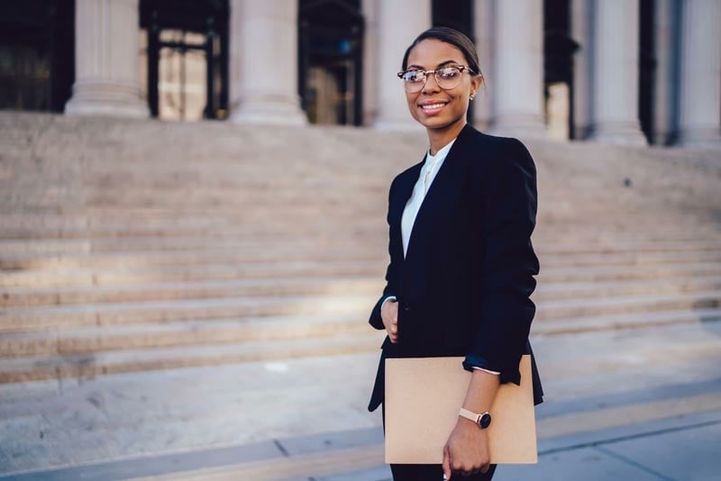 A person in business attire stands outside a building with columns and steps, holding a folder and smiling at the camera.