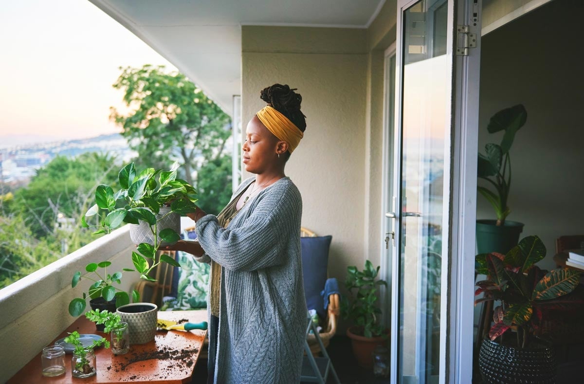 A woman stands on a balcony tending to potted plants, with gardening tools and soil on a table beside her. The balcony overlooks a green, tree-filled landscape.