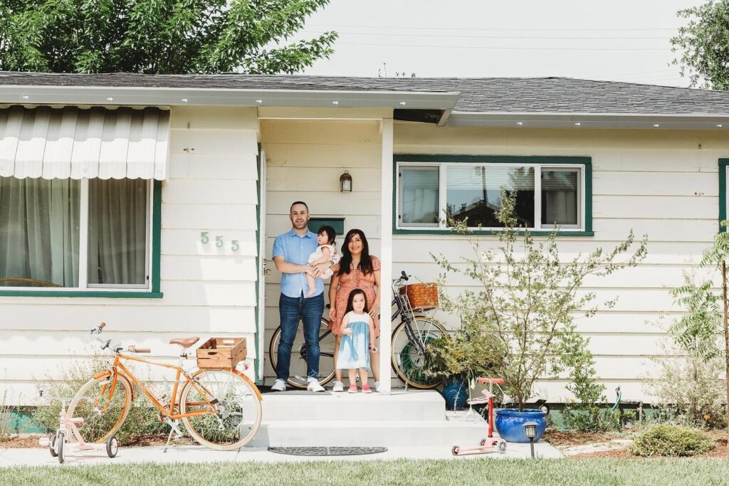 A family of four stands on the front steps of a single-story house with two bicycles, a child's scooter, and toys in the yard.