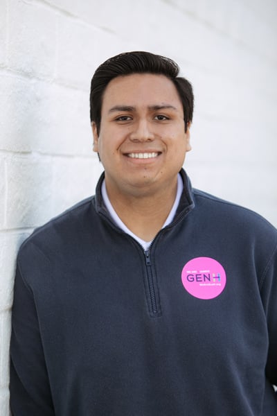 A person with short dark hair wearing a navy zip-up sweater and a pink "GEN H" sticker stands against a white brick wall, smiling at the camera.