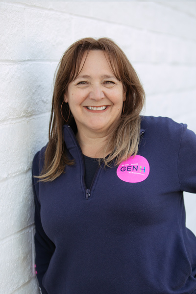Woman with brown hair wearing a navy sweater and a pink "GEN H" sticker, smiling while leaning against a white brick wall.