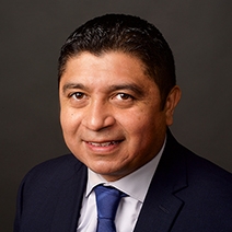 A man with short dark hair in a navy suit, white shirt, and blue tie poses against a plain dark background.