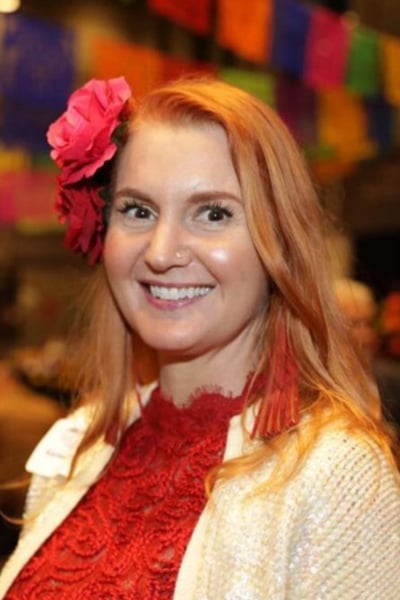 A woman with long red hair, wearing a red lace top, cream sweater, red flower in her hair, and red earrings, smiles at the camera. Colorful banners hang in the blurred background.