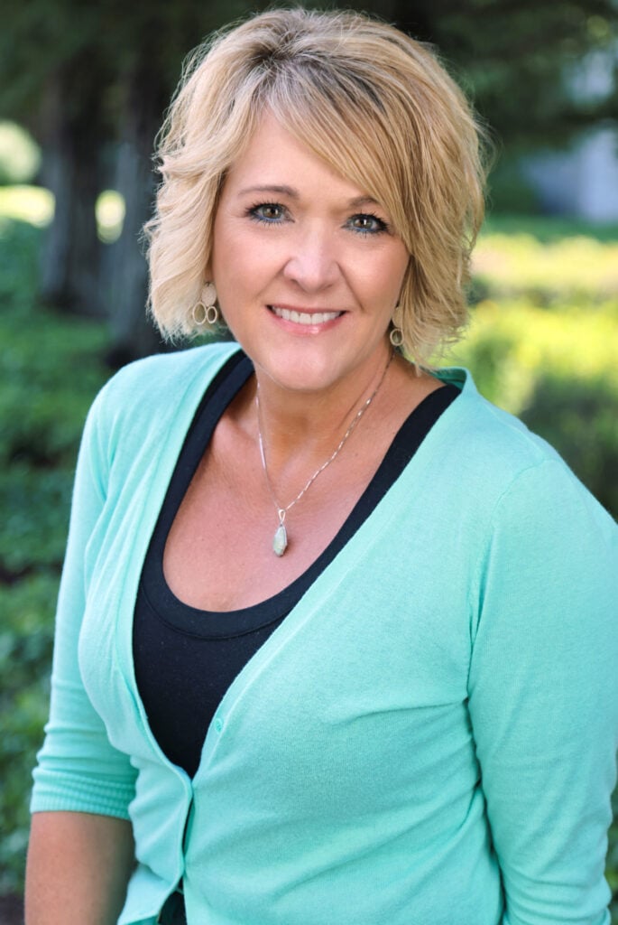 A woman with short blonde hair wearing a mint green cardigan and a necklace is smiling at the camera outdoors, with greenery in the background.