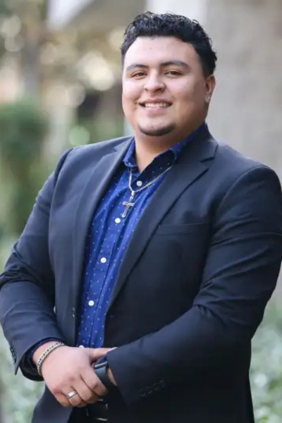 A man in a navy suit and blue shirt stands outdoors, smiling with his arms crossed in front of him.