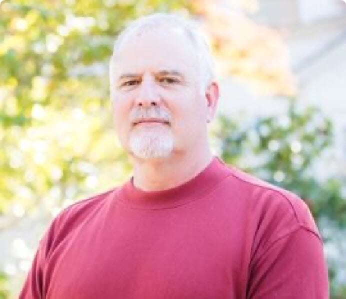 A man with short white hair and a goatee wearing a red sweatshirt stands outdoors with blurred greenery in the background.