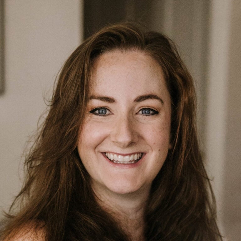 A woman with long brown hair smiles at the camera, standing indoors with a blurred background.