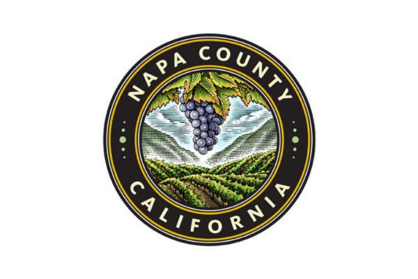 Circular seal for Napa County, California, featuring a bunch of grapes, green vineyards, hills, and a sky in the center, with text reading "Napa County California" around the border.