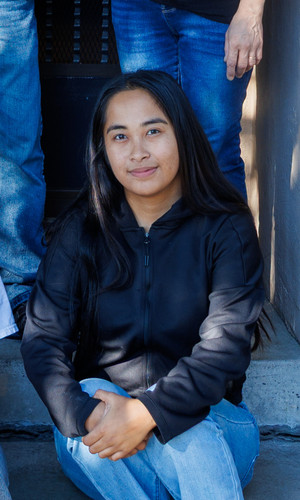 A young woman with long dark hair, wearing a black jacket and blue jeans, sits on concrete steps with her arms resting on her knees. Two people are partially visible beside her.