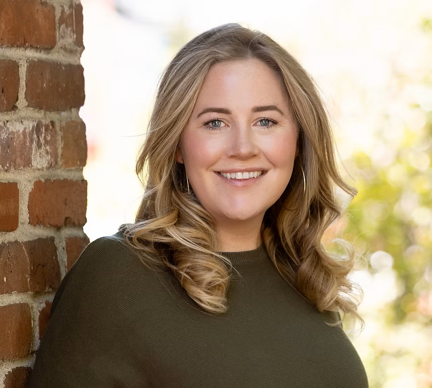 A woman with wavy blonde hair wearing a dark green top smiles while standing next to a brick wall outdoors.