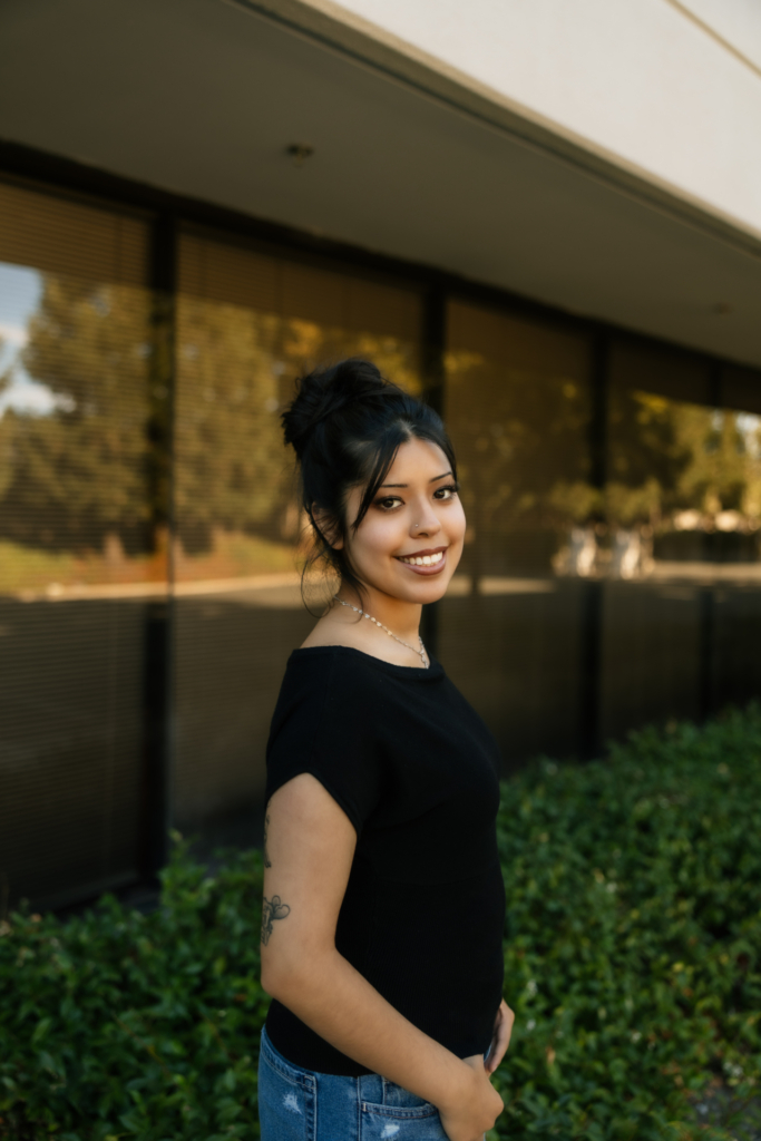 A smiling young woman wearing a black shirt stands in front of a modern building with large windows and a hedge.